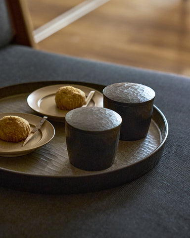 Two black wooden cups with textured lids by Ryuiji Mitani with pastries on wooden plates on a dark tray with a blurred background