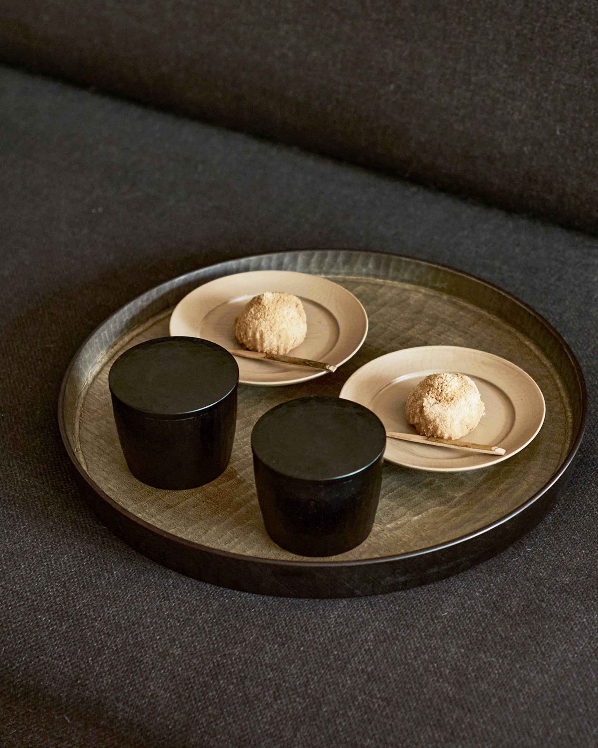 Two black wooden cups and two beige plates with food on a wooden tray all made by Ryuji mitani on a dark gray surface