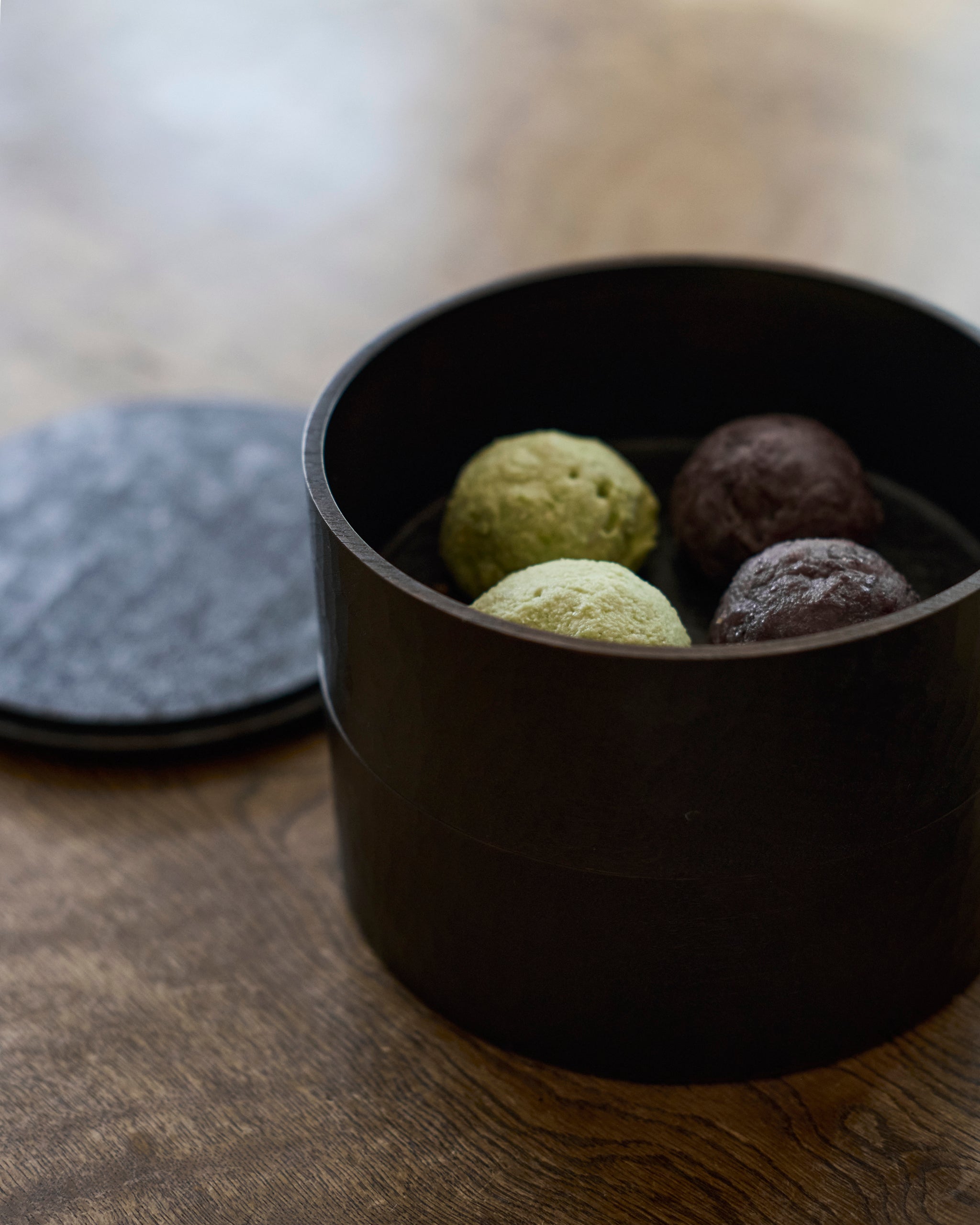 Opened wooden two-tiered box by Ryuji Mitani with green and purple spherical treats on a wooden surface