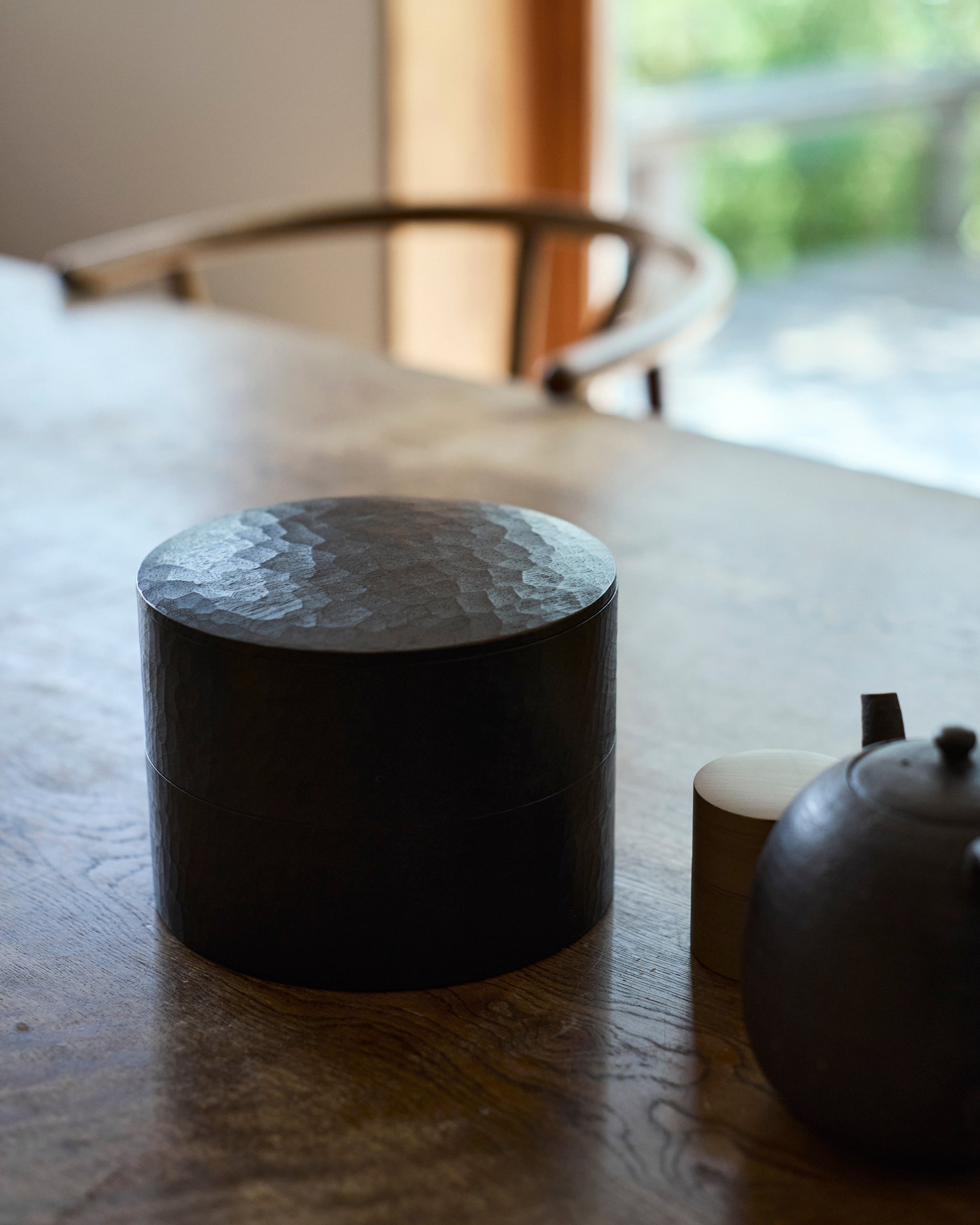 Cylindrical wooden two-tiered box by Ryuji Mitani with textured top on a wooden surface, with a teapot and small cylindrical box in the background.