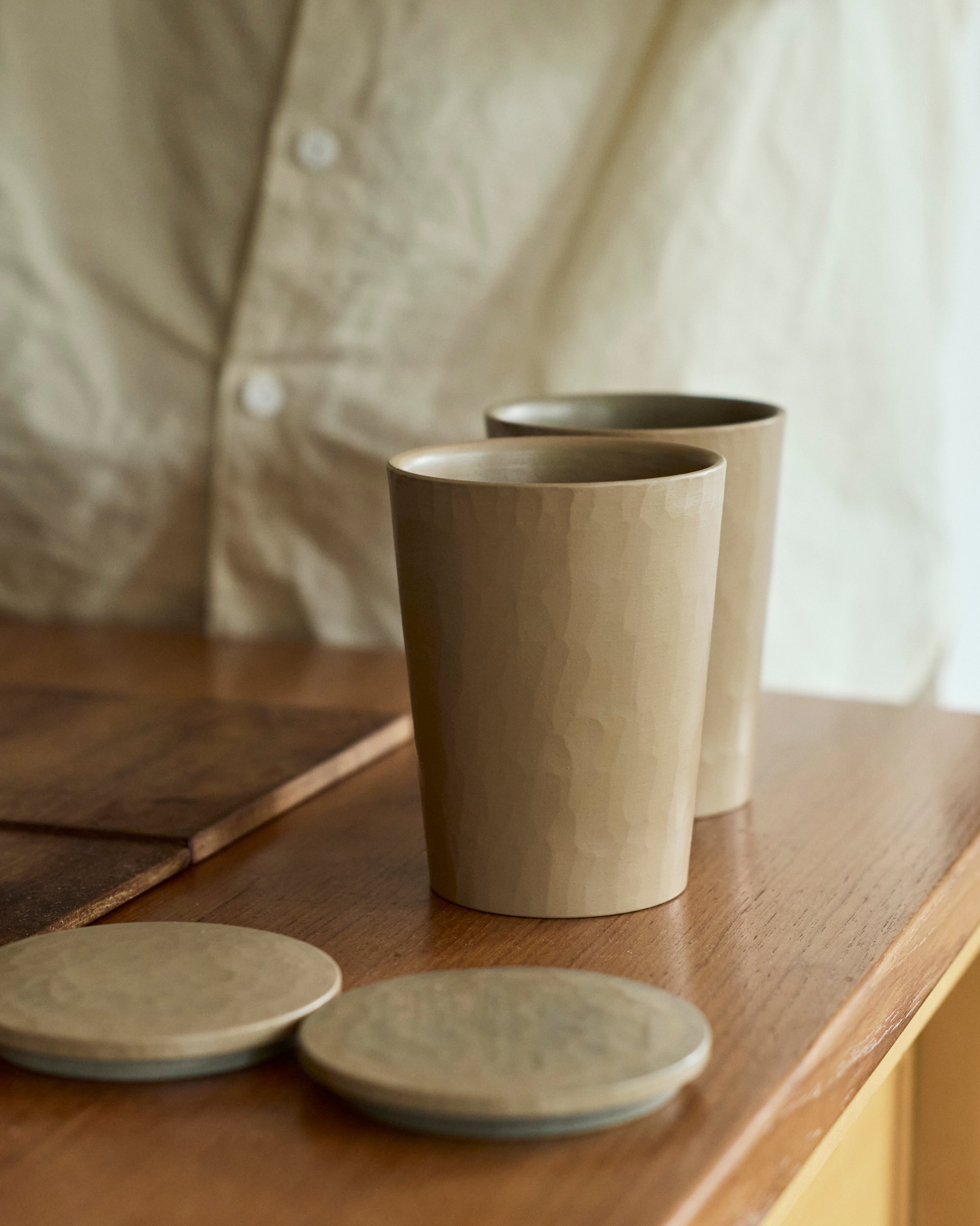 Two wooden cups and their lids by Ryuji Mitani on a wooden surface with a blurred person in the background.