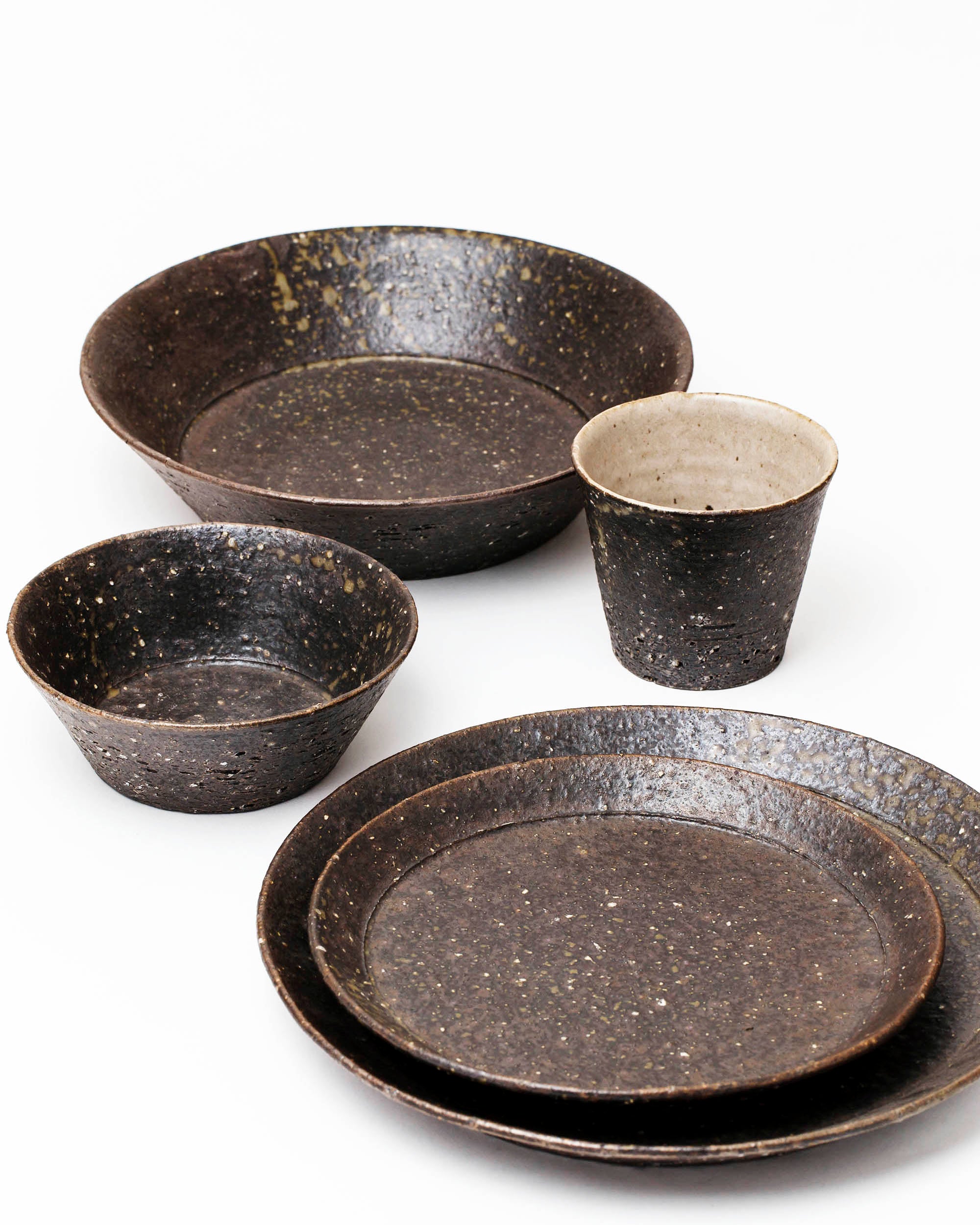 Many brown textured plates and bowl against a white background. On the left there is a cup with a beige interior.