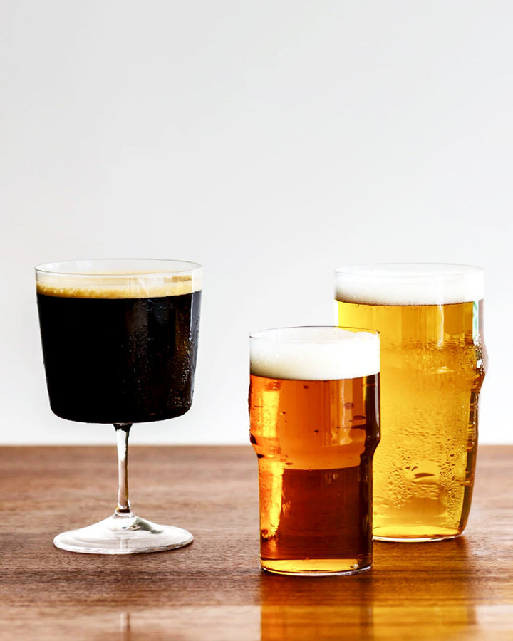 Three time and style glasses side by side on a wood table. Raisin glass is on the left with dark poured beer inside.