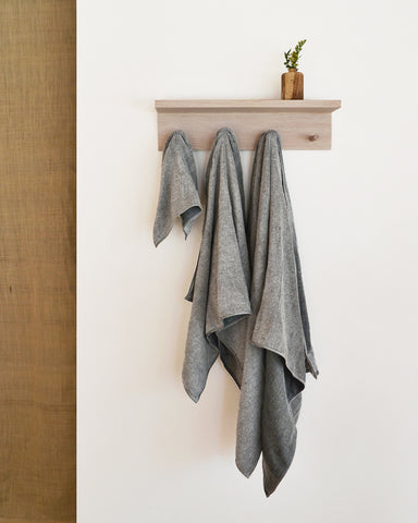 Body, hand, and face zen charcoal towels are hanging on a white oak hanger on a white wall. A wood vase is placed on top of the hanger.