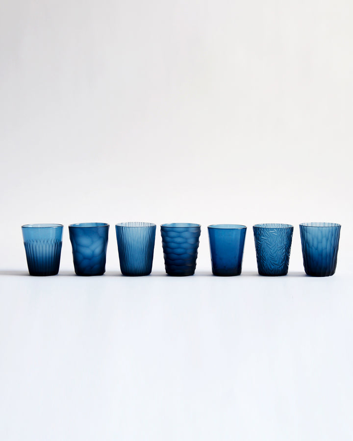 A row of seven cups from factory zoomer's reclaimed blue 'nature's diary' glass set. Each cup features a unique carved pattern.