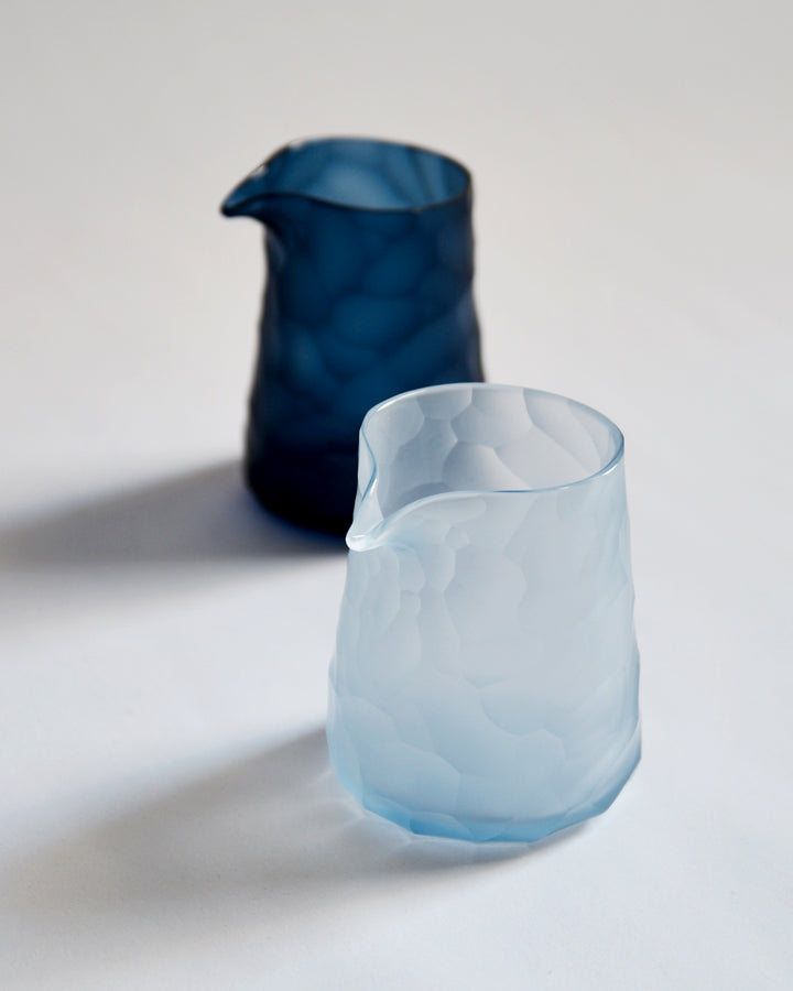 Light blue reclaimed blue one lipped pitcher is placed in front and the dark blue pitcher is in the back. 