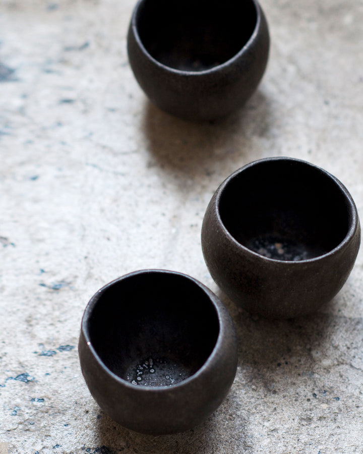 Three round black ceramic pod cup handcrafted by Keisuke Iwata against concretebackground