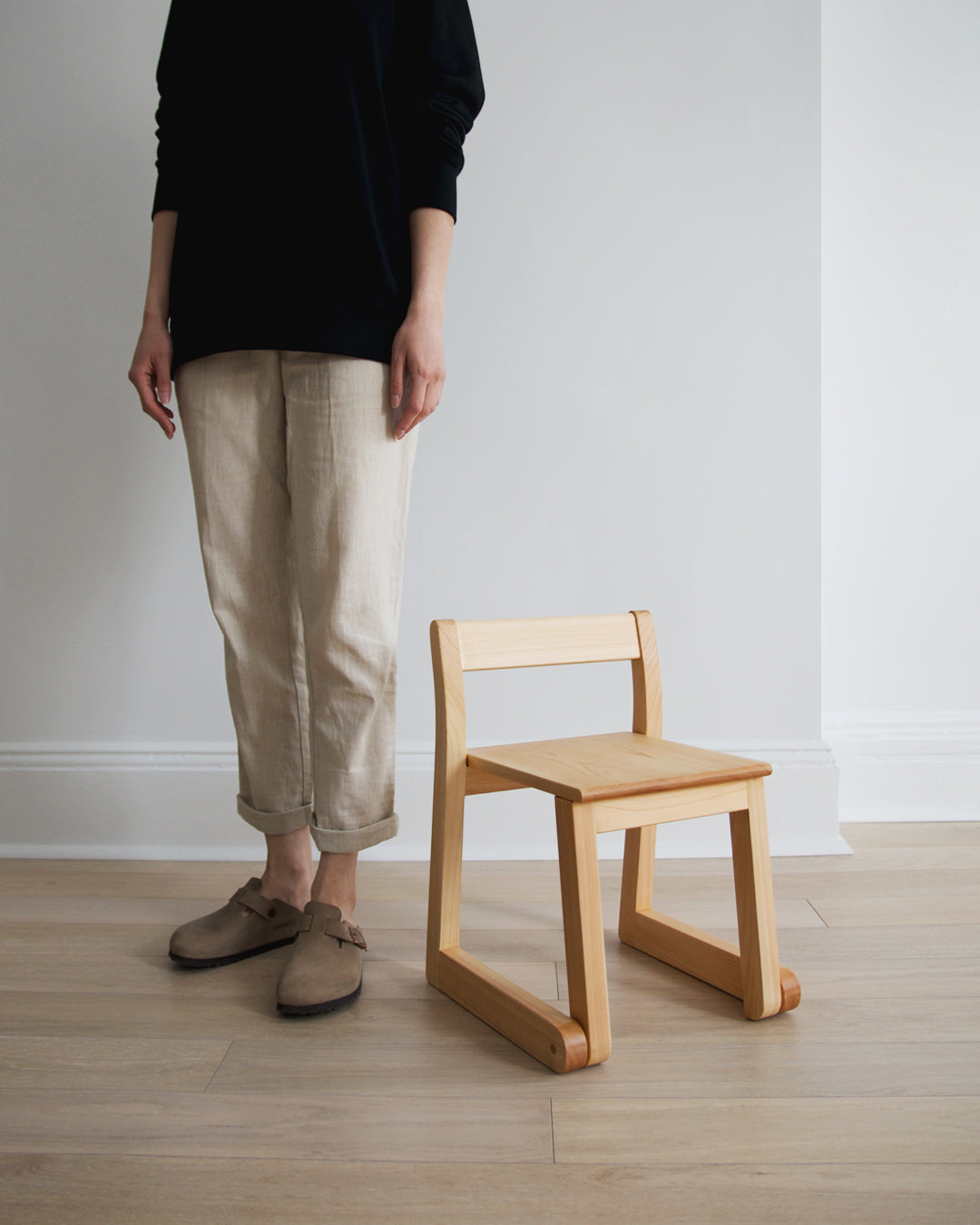A woman in black t-shirt and beige pant standing next to the 5 year old school chair by makoto koizumi.