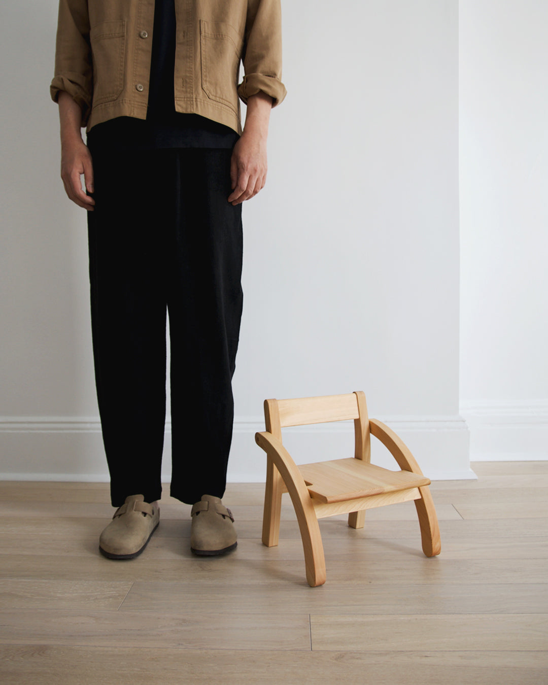 A man in all black attire with khaki clogs standing next to the 1 year old school chair by makoto koizumi.