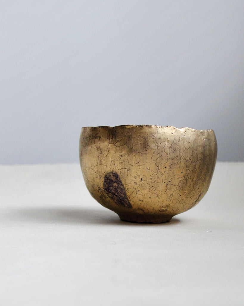 Gold Chawan III on a white table with gray background.