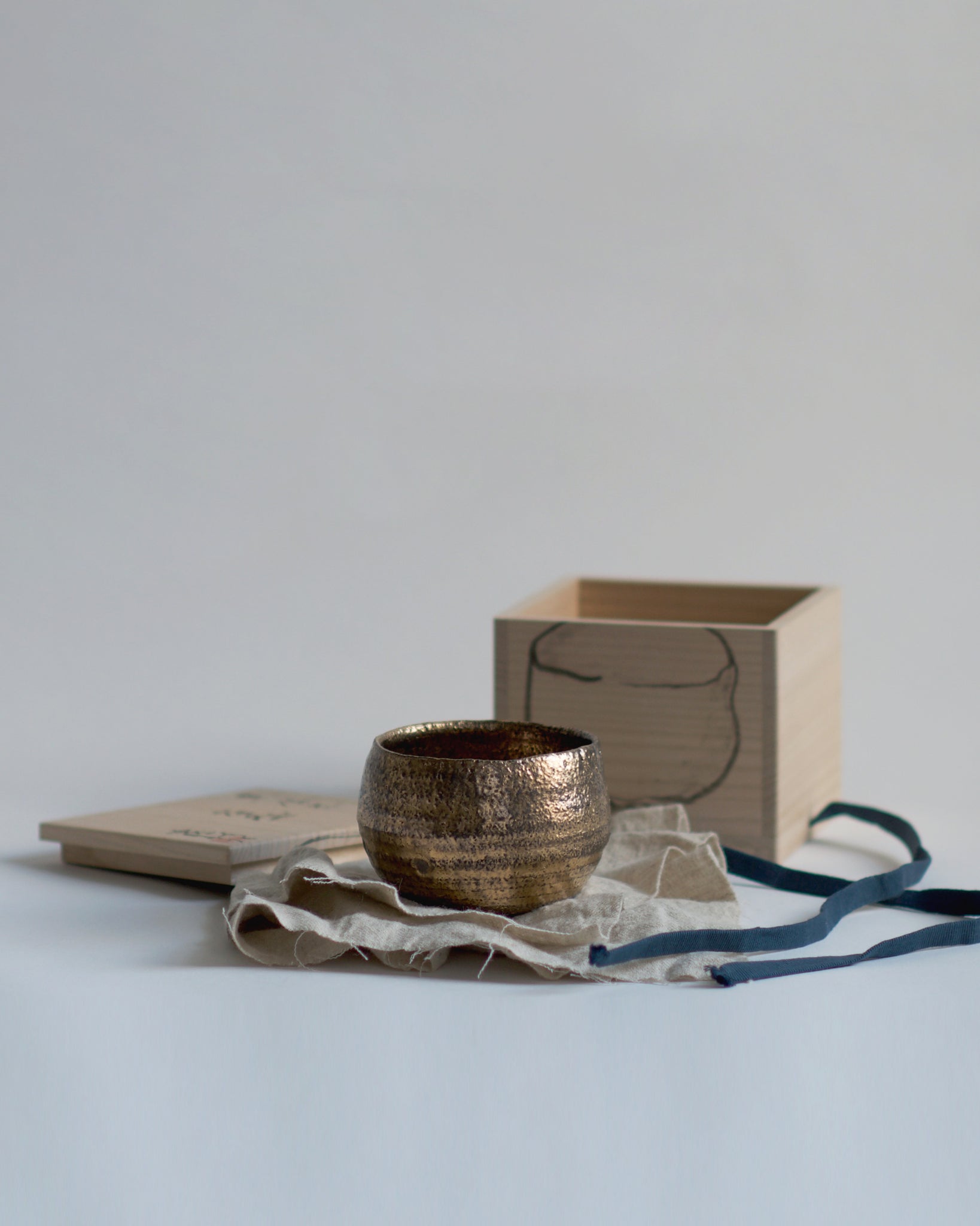 Gold Chawan V on top of a wrapping cloth. Wooden box calligraphed by Masanobu Ando in the background.