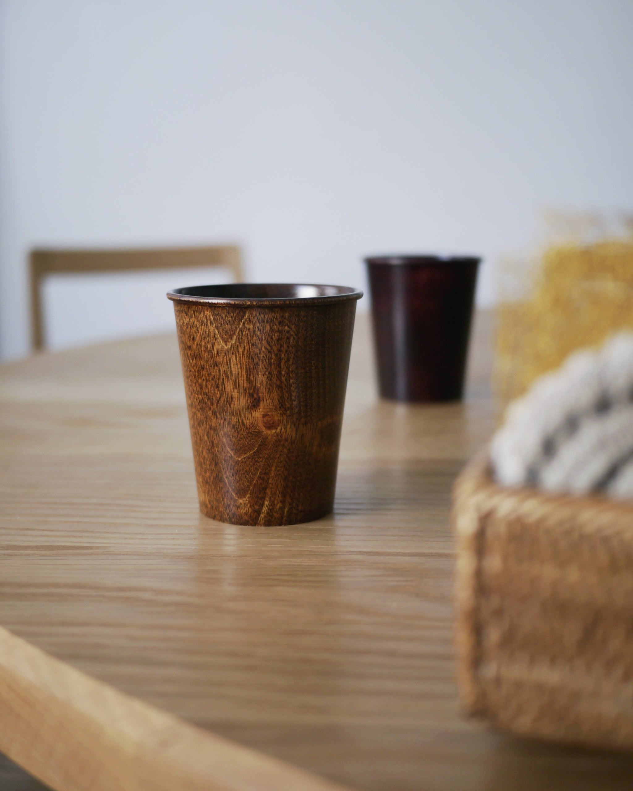 Brown urushi wood cup on a wood dining table with a red urushi wood cup blurry in the background
