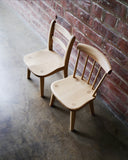 An angled view of Ladderback and Spindle Back Children's Chairs standing side by side on a cement floor with brick wall.