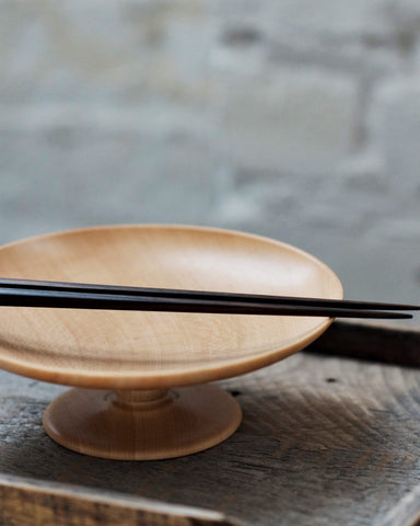 The footed plate is on top of a dark wood tray, and a pair of ebony wood chopsticks are placed on top of the footed tray.
