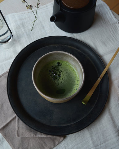 In-situation image of the uchugin silver matcha bowl with matcha inside. The matcha bowl is placed on top of a blackened brass tray, that is placed on top of natural tone linens. There is a bamboo matcha whisk besides the matcha bowl, and a cast iron teapot on the top corner of the image.