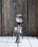 Sori yanagi stainless steel serving spoon and fork are placed in a glass jar, and placed on a white wood surface.